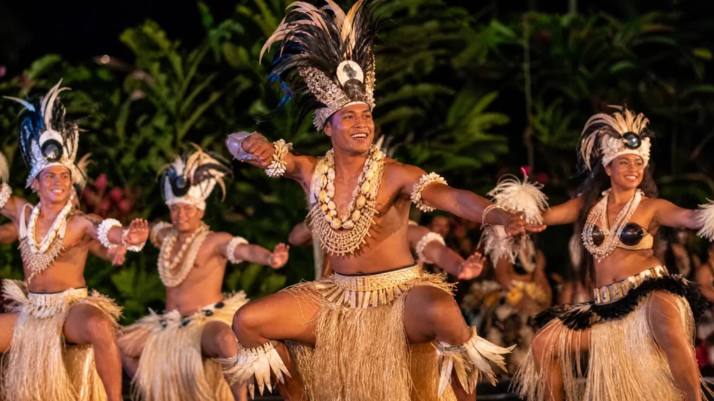 A professional editorial photograph of traditional Cook Islands dancers in intricate Polynesian costumes made of fibers and shells, performing an energetic rhythmic dance on a stage under warm evening lighting.