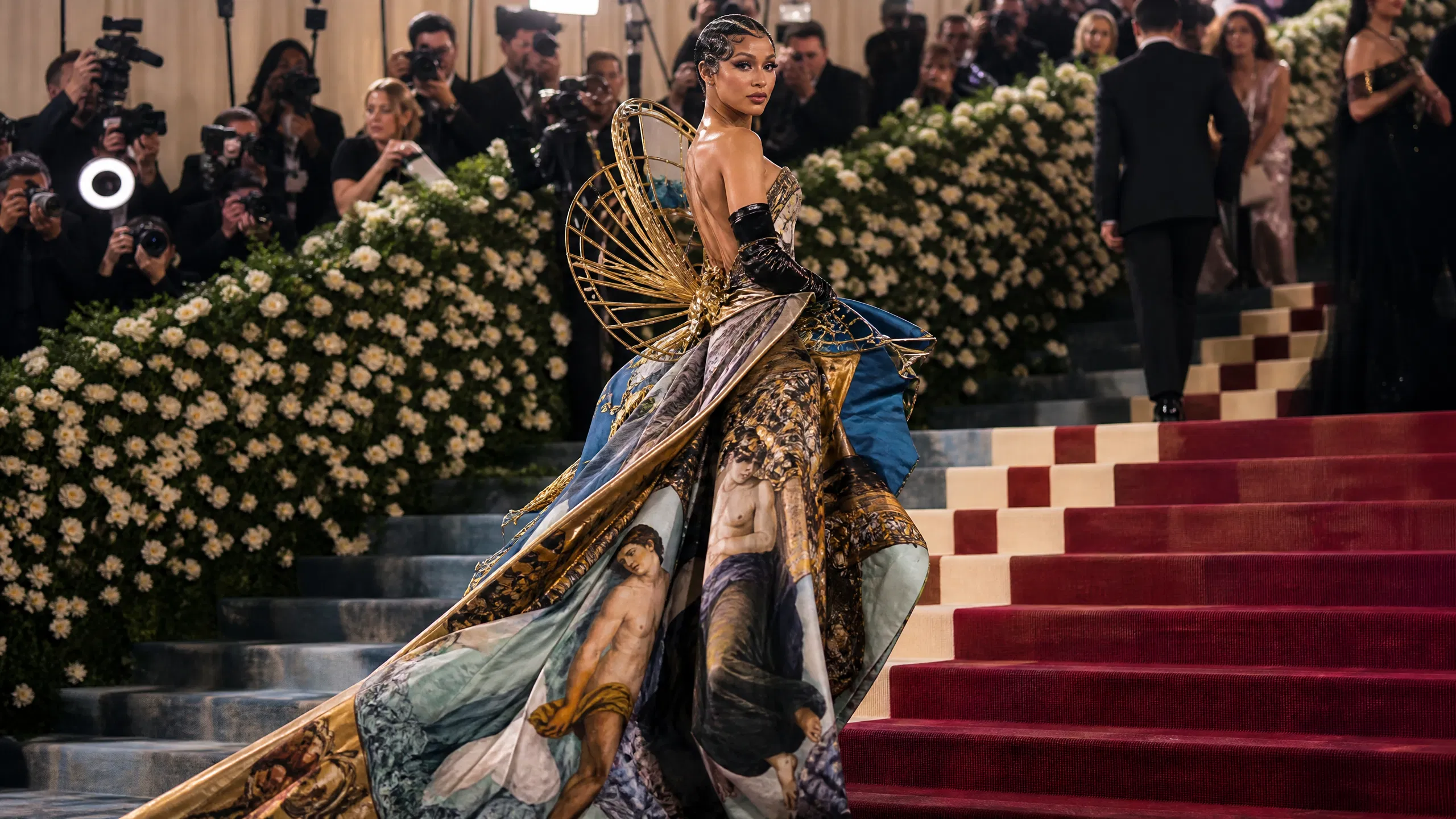 A professional editorial photograph of a celebrity on the grand red carpet steps of the Metropolitan Museum of Art for the 2026 Met Gala. The 'Costume Art' theme is represented by an avant-garde gown blending classical artistic motifs with modern structural design, surrounded by paparazzi and theatrical lighting.1787069236840 na1fn L2hvbWUvdWJ1bnR1L21ldF9nYWxhXzIwMjY
