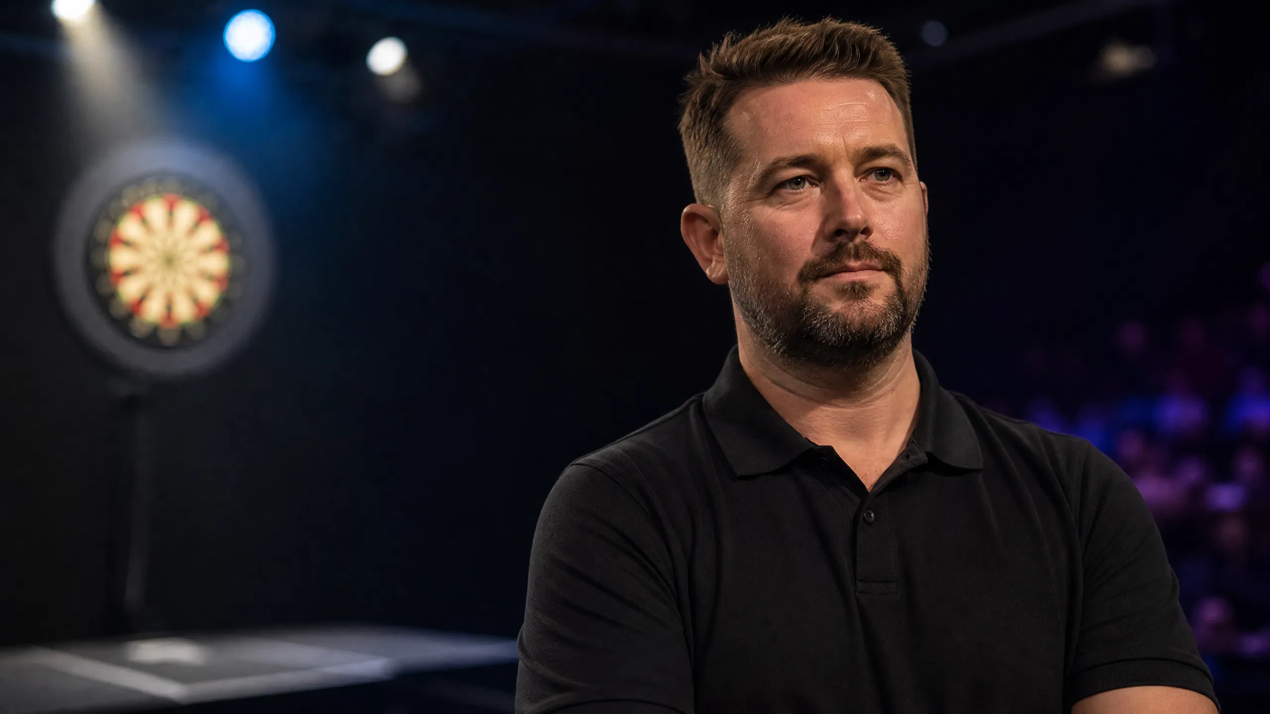 Editorial portrait of a professional darts player in a softly lit venue with a blurred dartboard behind him, representing mental-health resilience and advocacy.